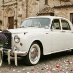 Voiture de mariage vintage décorée d'un balai en lavande et eucalyptus avec rubans de satin ivoire, garée sur une place pavée provençale