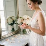 Mariée en robe de dentelle ivoire qui insère une lentille de contact devant un miroir vintage dans une suite nuptiale décorée de pivoines