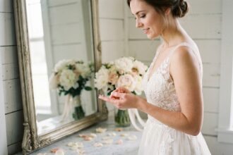 Mariée en robe de dentelle ivoire qui insère une lentille de contact devant un miroir vintage dans une suite nuptiale décorée de pivoines